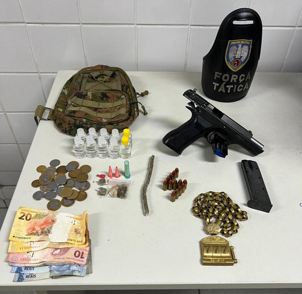 Sobre mesa materiais apreendidos pela Polícia Militar, uma pistola 9 mm, 10 frascos de loló, quatro porções de maconha, uma pedra de crack, três pinos de cocaína, R$ 91,15 em dinheiro, um carregador compatível com a pistola apreendida e seis munições, além de um cordão dourado com o pingente "777".
