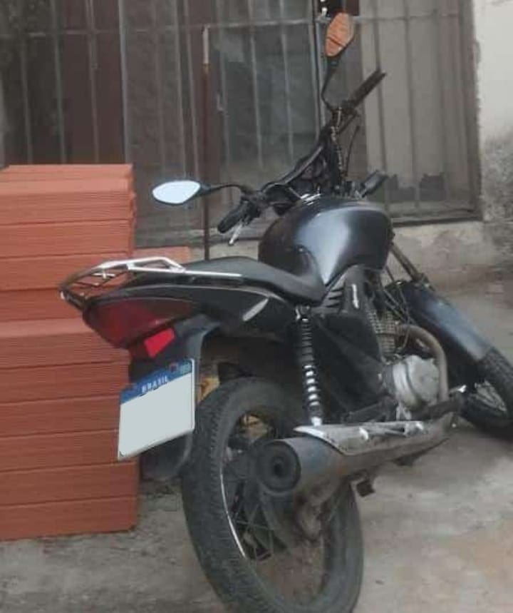 Imagem de uma moto Honda CG 150 estacionada ao lado de uma pilha de blocos.