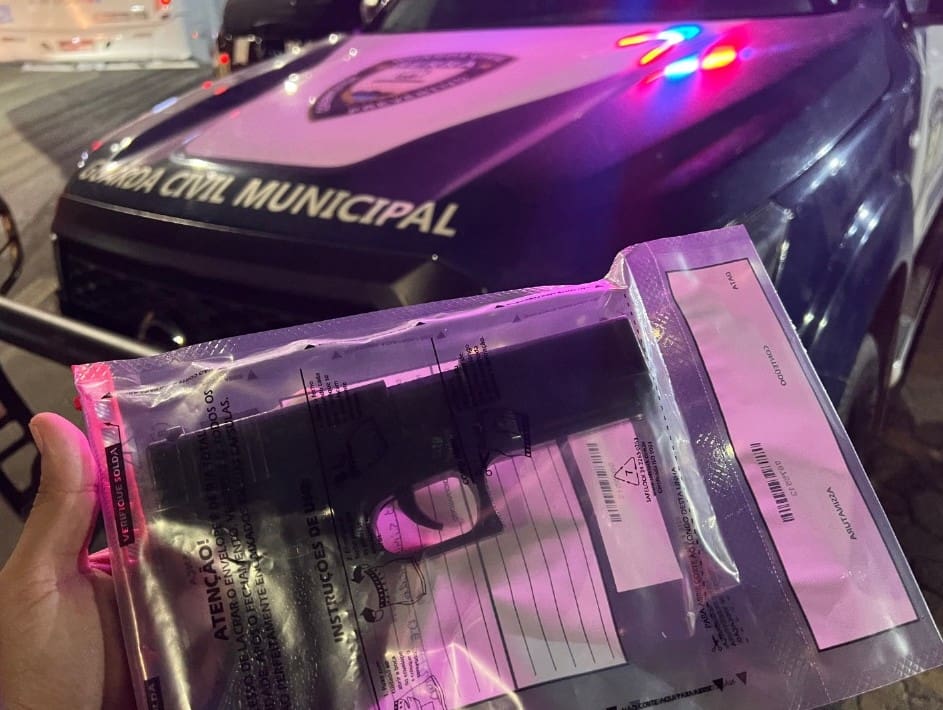 Um agente segurando um simulacro de arma de fogo apreendido com os assaltantes, em frente a uma viatura da Guarda da Serra.