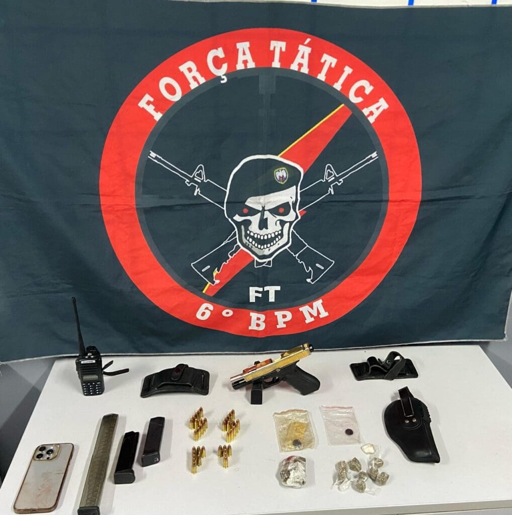 Uma sala com uma bandeira da Força Tática do 6º Batalhão, após uma ocorrência envolvendo traficantes, sobre uma mesa uma pistola, três carregadores, munições e drogas. 