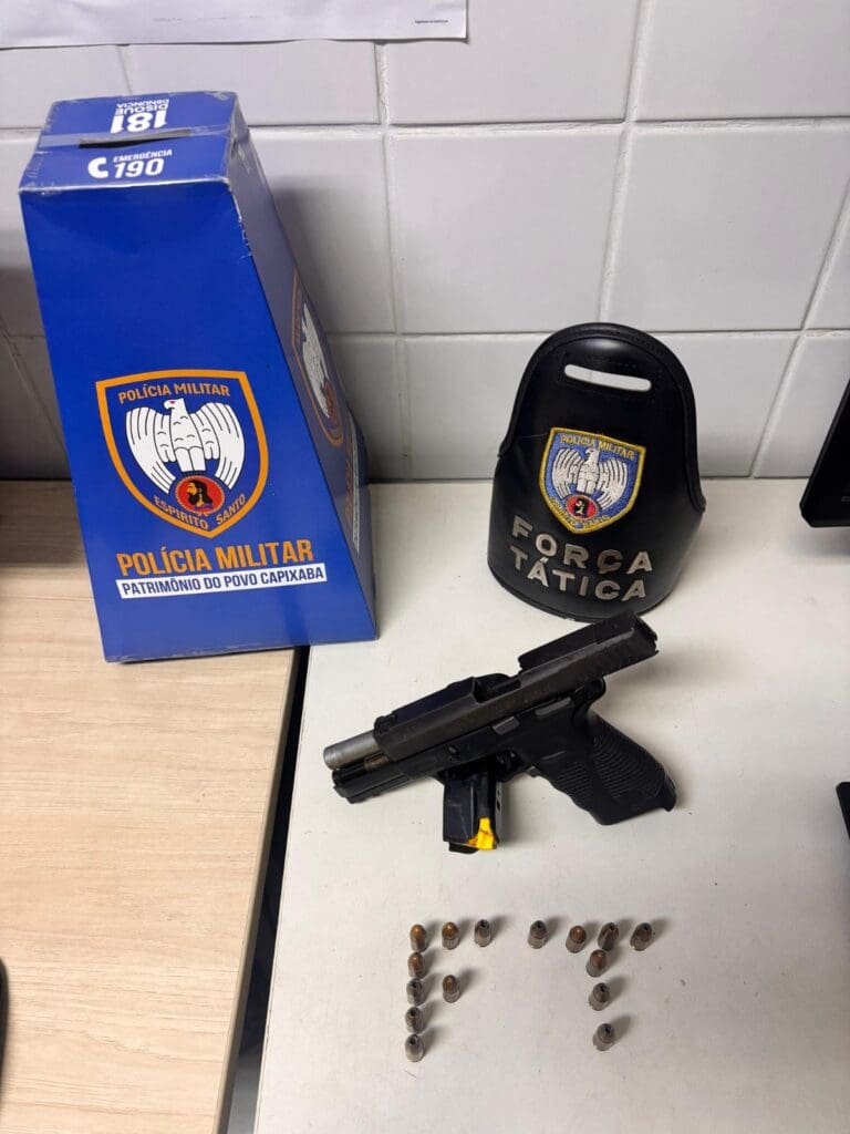 Imagem de uma pistola, 15 munições, um totem de papelão de divulgação da PMES e uma braçadeira de identificação da Força Tática sobre uma mesa de uma delegacia.