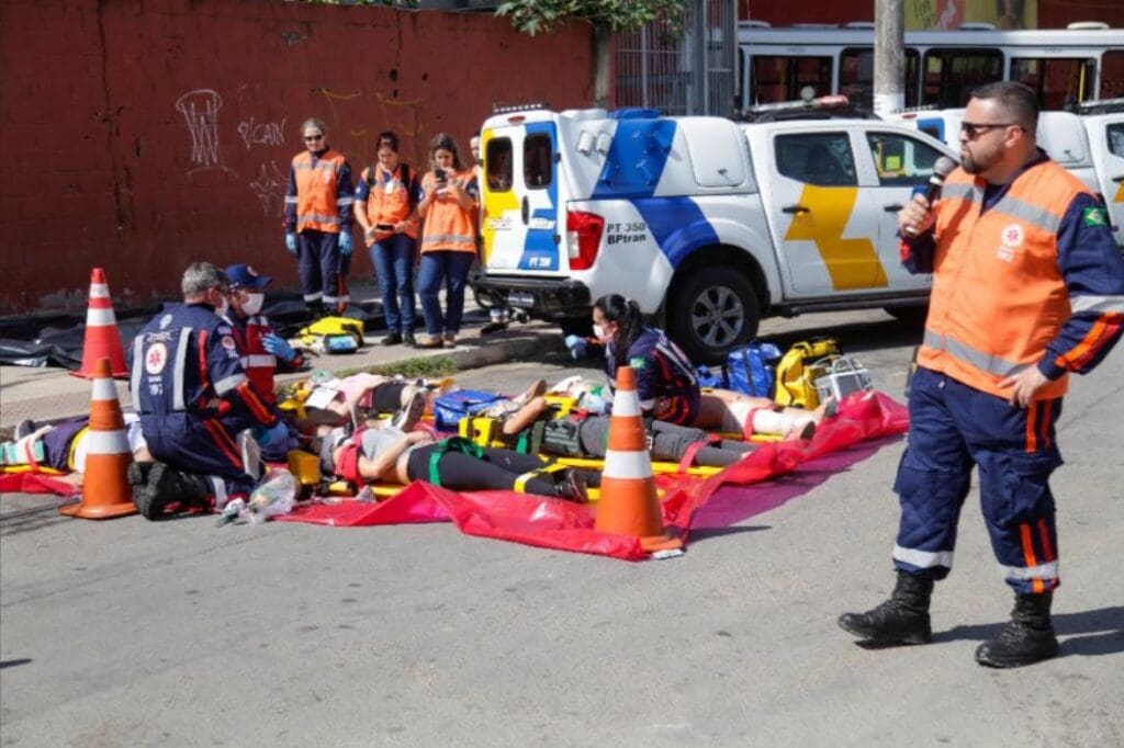 Imagem de um socorrista do SAMU segurando um microfone, enquanto outros socorristas simulando atendimento de vítimas que estão deitadas na rua.