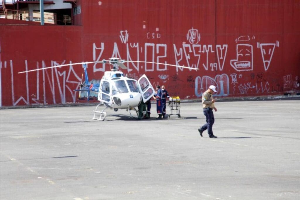 Imagem de uma simulação de um acidente: Um helicóptero pousado em um estacionamento com socorristas colocando uma maca na aeronave. 