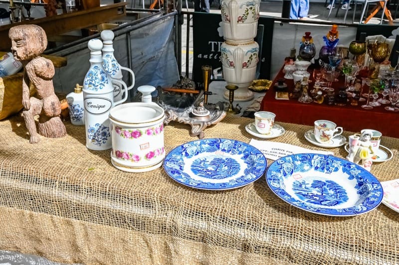 Imagem de diversos itens antigos expostos sobre uma mesa de um comerciante (esculturas, louças de porcelana entre outros itens), na Feira de Antiguidade da Rua da Lama em Jardim da Penha.