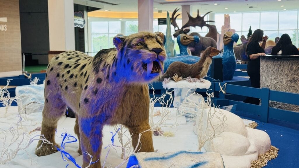Imagem da Exposição “Gigantes da Era do Gelo”, no saguão de um shopping, com bonecos gigantes de um tigre dentes de sabre e outros animais pré-históricos, no fundo. 