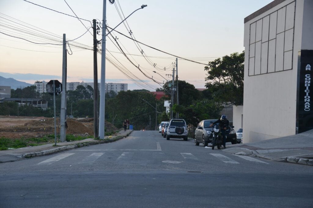 Imagem da entrada de uma rua com um terreno ao lado esquerdo e carros estacionados do lado direito da rua.