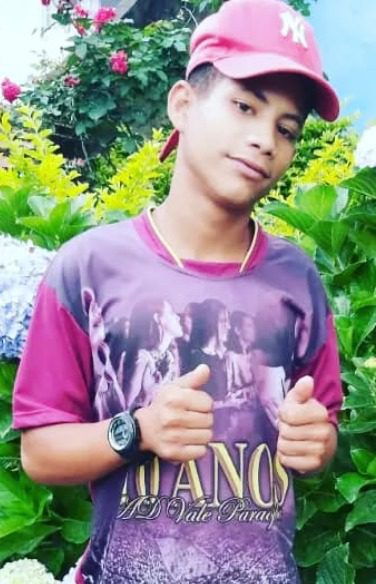 Imagem de um jovem (Frederico Gonçalves dos Santos), com as duas mãos fazendo sinal de positivo e no fundo um jardim florido.