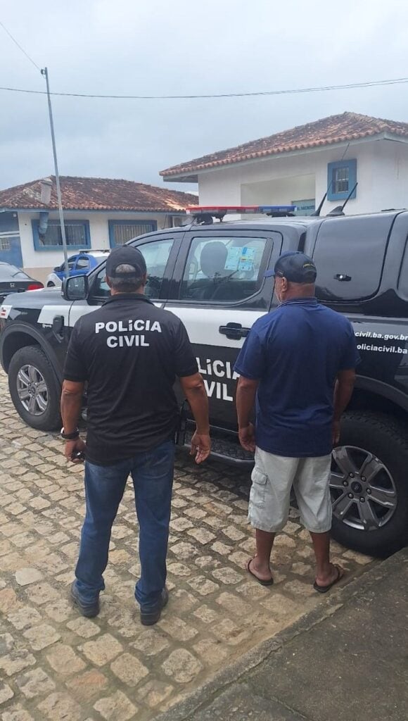 Foto de um policial civil ao lado de um homem, em um patio de uma delegacia com uma viatura da Polícia Civil.