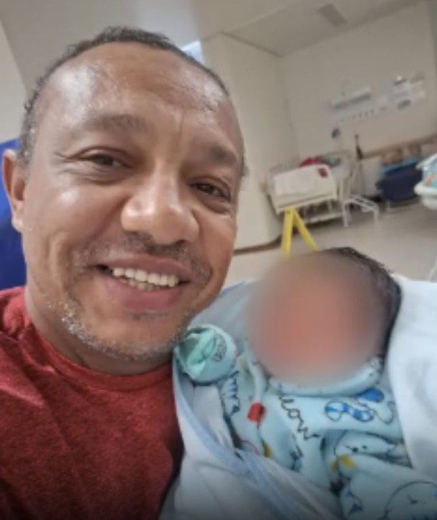 Foto em destaque de um homem segurando um bebê e no fundo uma viatura utilizada para transportar corpos da Polícia Cientifica.