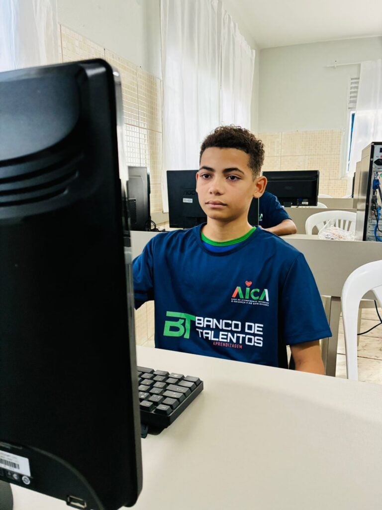 Um adolescente usando a camiseta do Banco de Talentos Aprendizagem da Rede AICA, sentado estudando usando um computador.