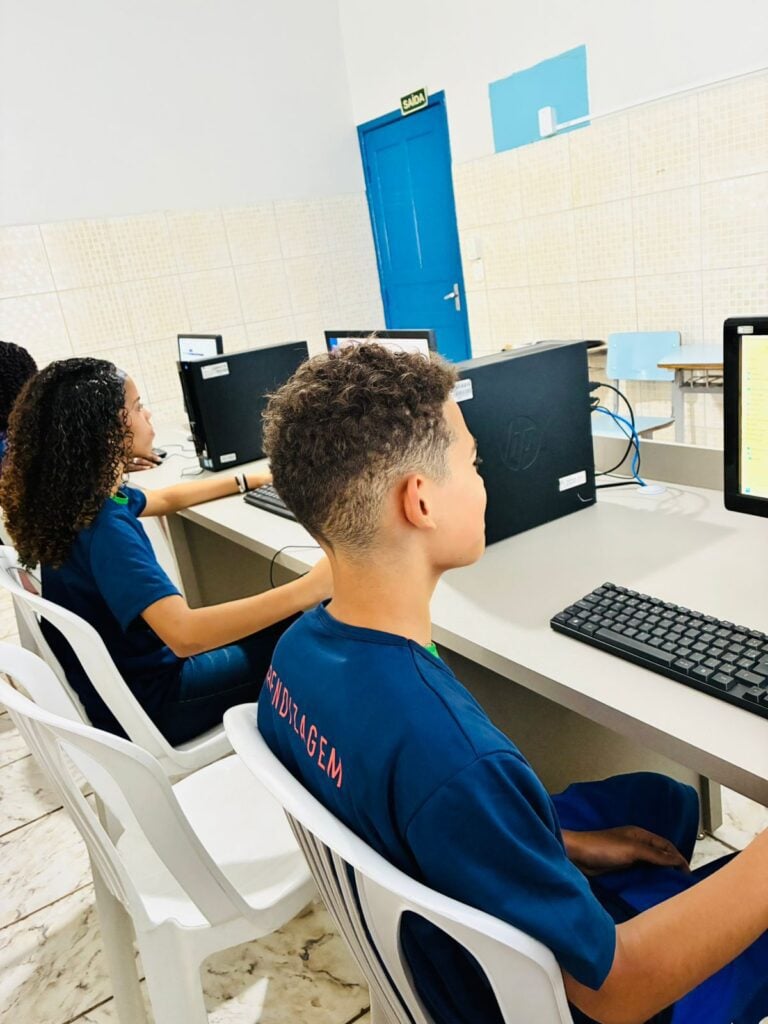 Três adolescentes usando a camiseta do Banco de Talentos Aprendizagem da Rede AICA, sentado estudando em computadores.