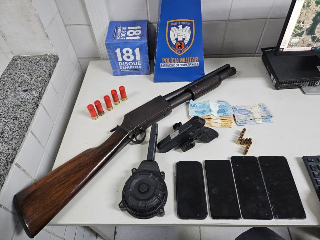 Sobre uma mesa armas apreendidas durante uma ocorrência realizada pela Polícia Militar em José de Anchieta. Uma espingarda calibre 12, cinco munições, uma pistola calibre 9mm, 12 munições, um carregador tipo caracol, quatro celulares e 196 reais em, espécie.