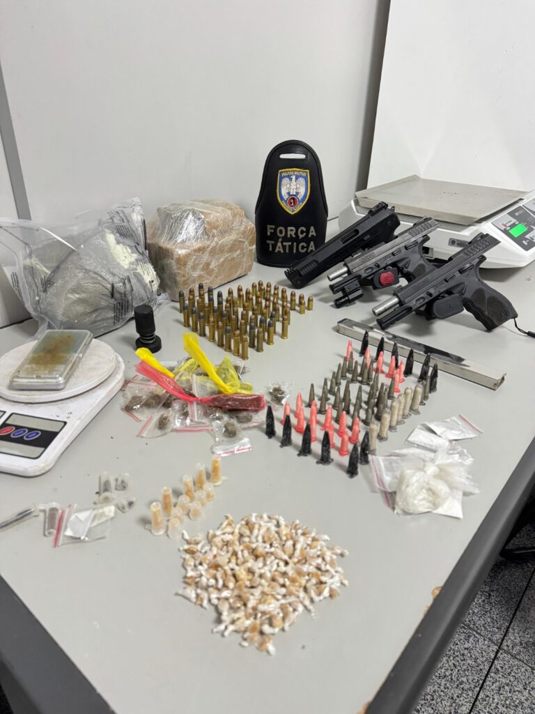 Imagem de vários materiais apreendidos pela Polícia Militar sobre uma mesa, duas pistolas, um simulacro de pistola, munições e diversos porções de drogas.