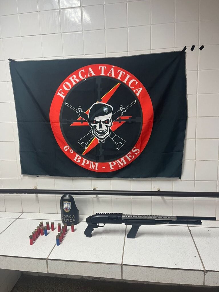 Imagem de apreensões da Polícia Militar em uma sala uma bandeira da Força Tática do 6º Batalhão (Uma caveira de boina com dois fuzis cruzados atras dela) e sobre uma bancada uma espingarda calibre 12 além de 17 munições do mesmo calibre.