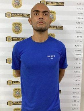 Imagem de um homem (Davidson Daniel de Carvalho Pereira, de 27 anos) alto com as mãos para trás, em frente a um banner da Polícia Civil.