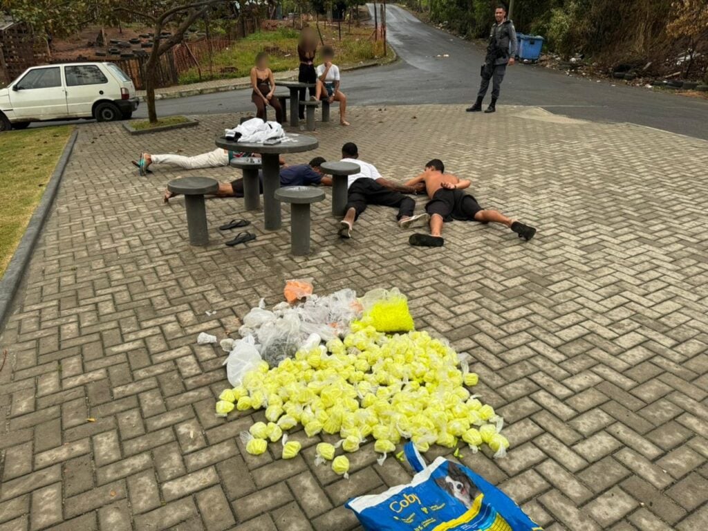 Em uma praça um policial militar, parado ao lado de três mulheres detidas, quatro homens deitados no chão e 6.721 pinos de cocaína espalhados durante uma ação da Polícia Militar.