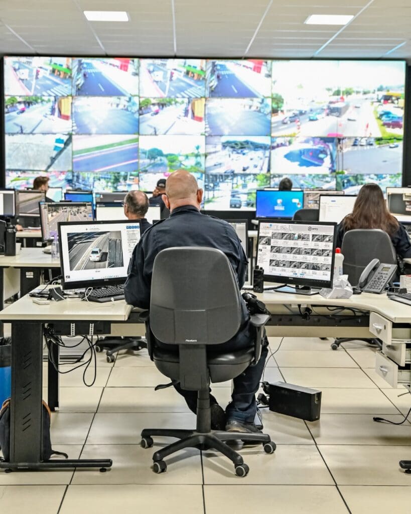 Imagem de uma sala de videomonitoramento com um telão exibindo diversas câmeras de segurança espalhadas pela cidade, e cerca de seis funcionários posicionados em frente a monitores de videomonitoramento.