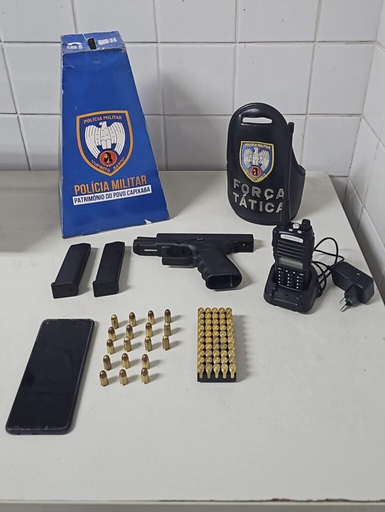 Sobre uma mesa materiais apreendidos pela PM, dois carregadores, uma pistola calibre .9 mm, um rádio comunicador, 68 munições calibre .9 mm, um celular.