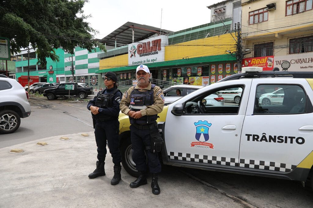 Imagem de dois agentes de trânsito próximos de uma viatura em um local com diversos comércios. 