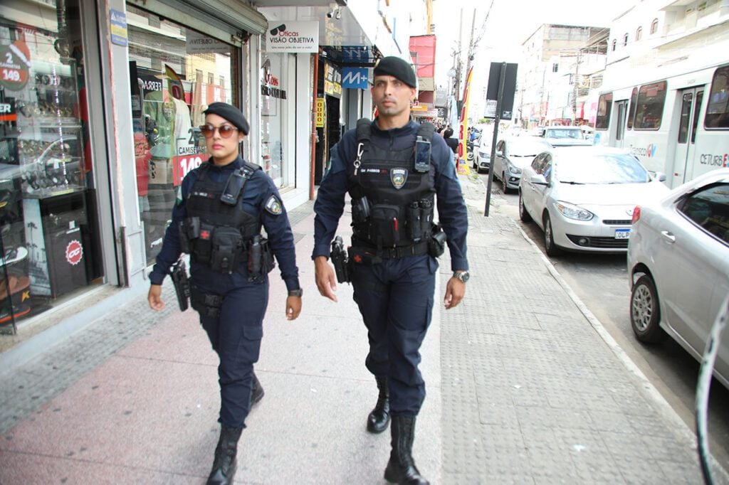 Imagem de dois agentes da Guarda de Cariacica andando em uma rua com diversos comércios.