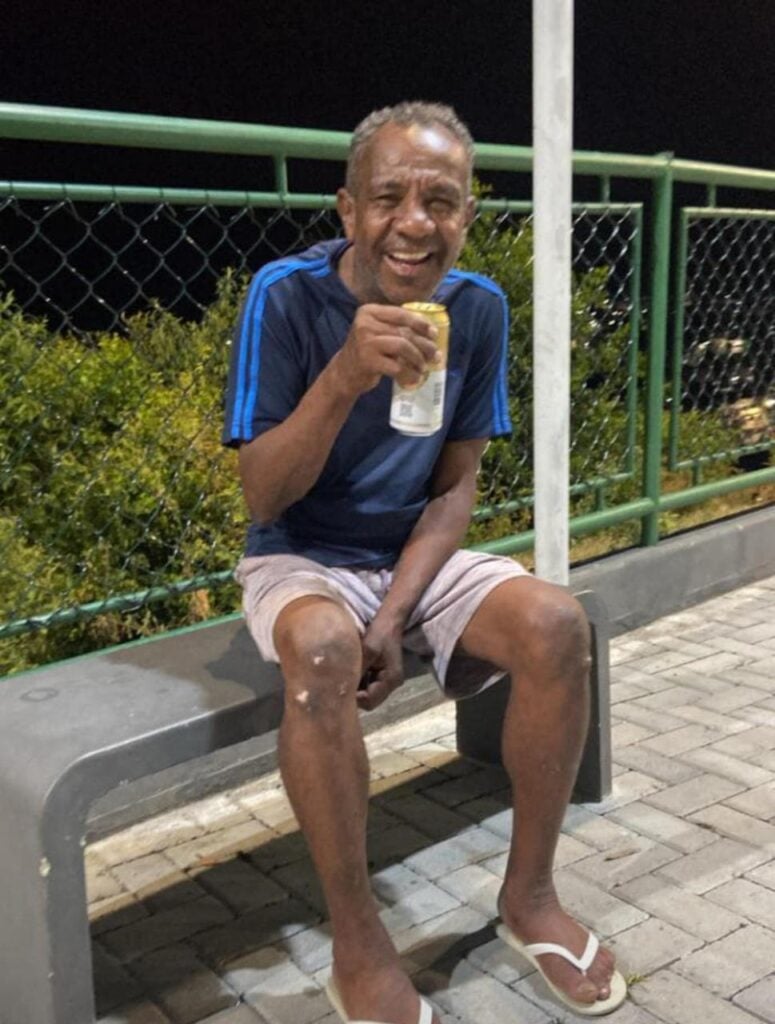 Um homem sorridente, sentado em um banco de uma praça segurando uma lata de cerveja.