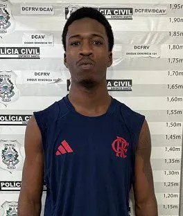 Imagem de um homem (Luiz Felipe Américo Constantino, de 21 anos), acusado de liderar uma quadrilha de furtos de moto, parado em frente a uma parede onde ha uma marcação de metros, usada pela polícia para realizar a fotografia e identificação de detidos.