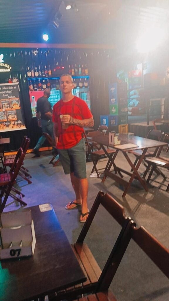 Um jovem (Gilvan Rodrigues de Oliveira Junior) segurando um copo com bebida, em um bar.