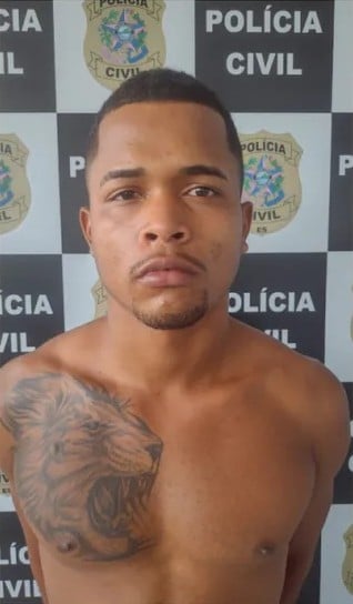 Foto de um homem (Rogério Silva Cardoso), com os braços para trás, em frente a uma parede com um banner da Polícia Civil.
