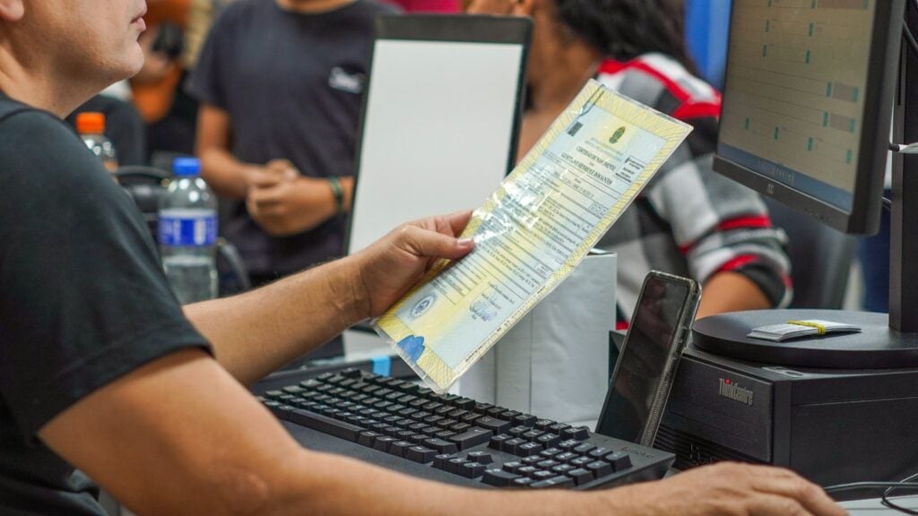 Imagem de uma mulher fazendo o atendimento durante o pedido de emissão da nova identidade, segurando um documento. E, no fundo, várias pessoas acompanham o processo.