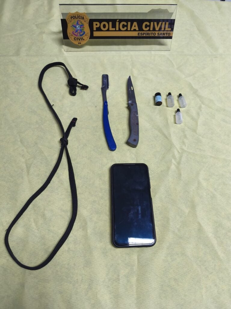 Sobre uma mesa alguns itens apreendidos pela Polícia Civil, uma faca, uma navalha, um cordão, quatro frascos com elementos quimicos e um aparelho celular,