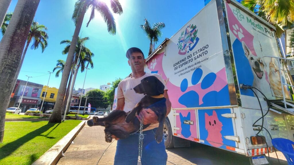 Um homem segurando um cachorro no colo ao lado de uma carreta estacionada em um praça.
